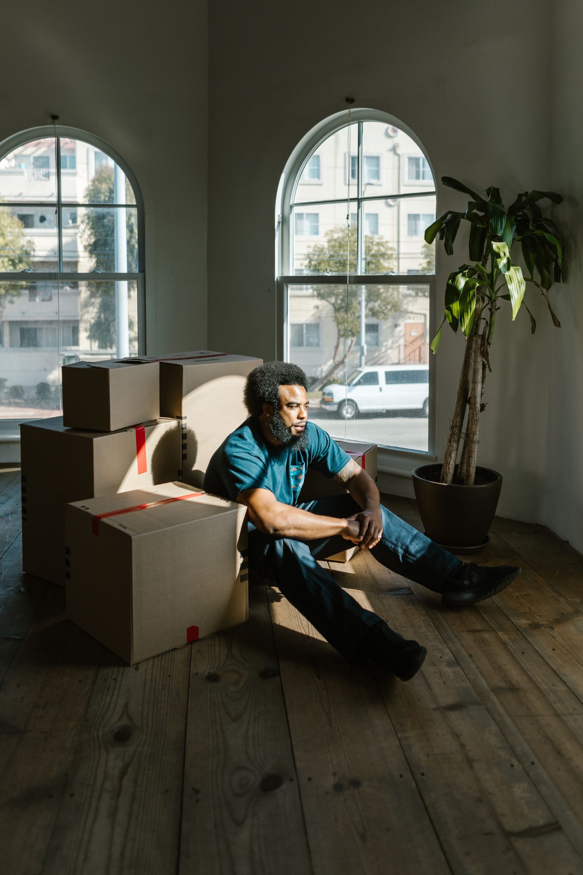 A man sits on the floor among stacked moving boxes in a bright room with tall arched windows, looking out at the street.