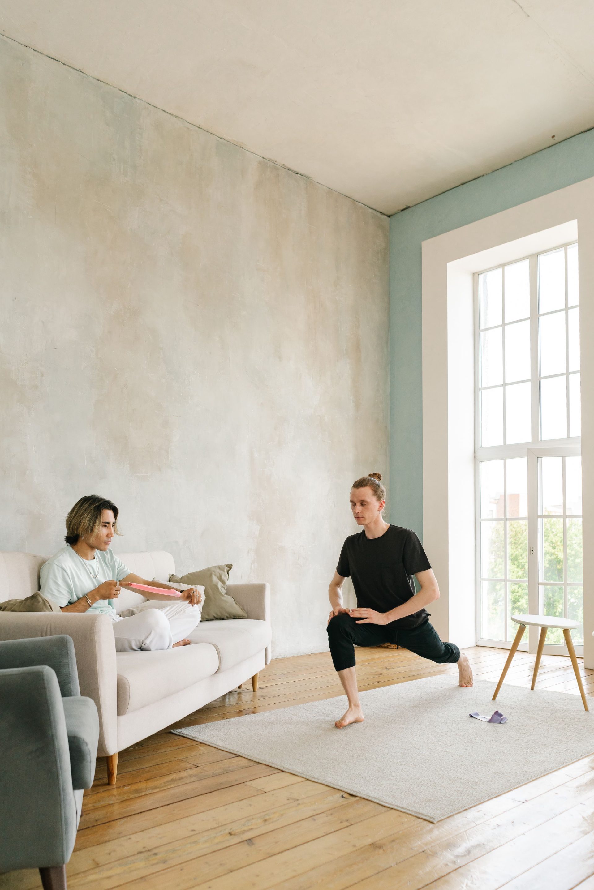 Two people in a sunlit living room: one holds a low lunge on a rug while the other sits on the sofa with a stretch band.